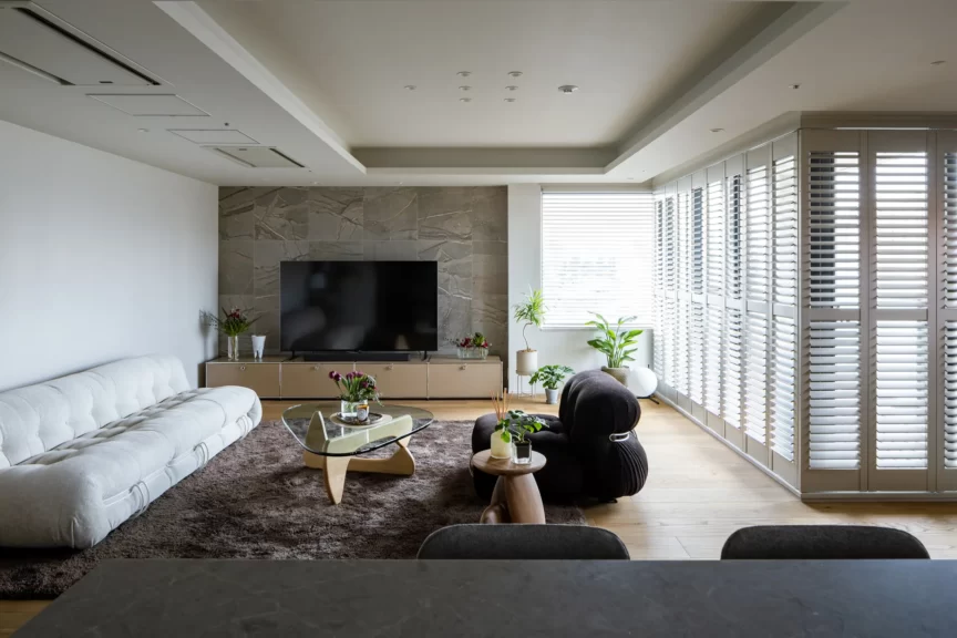 Open-plan living room with a wall-mounted TV on a marble accent wall, white sectional sofa, glass coffee table, and large windows with blinds.