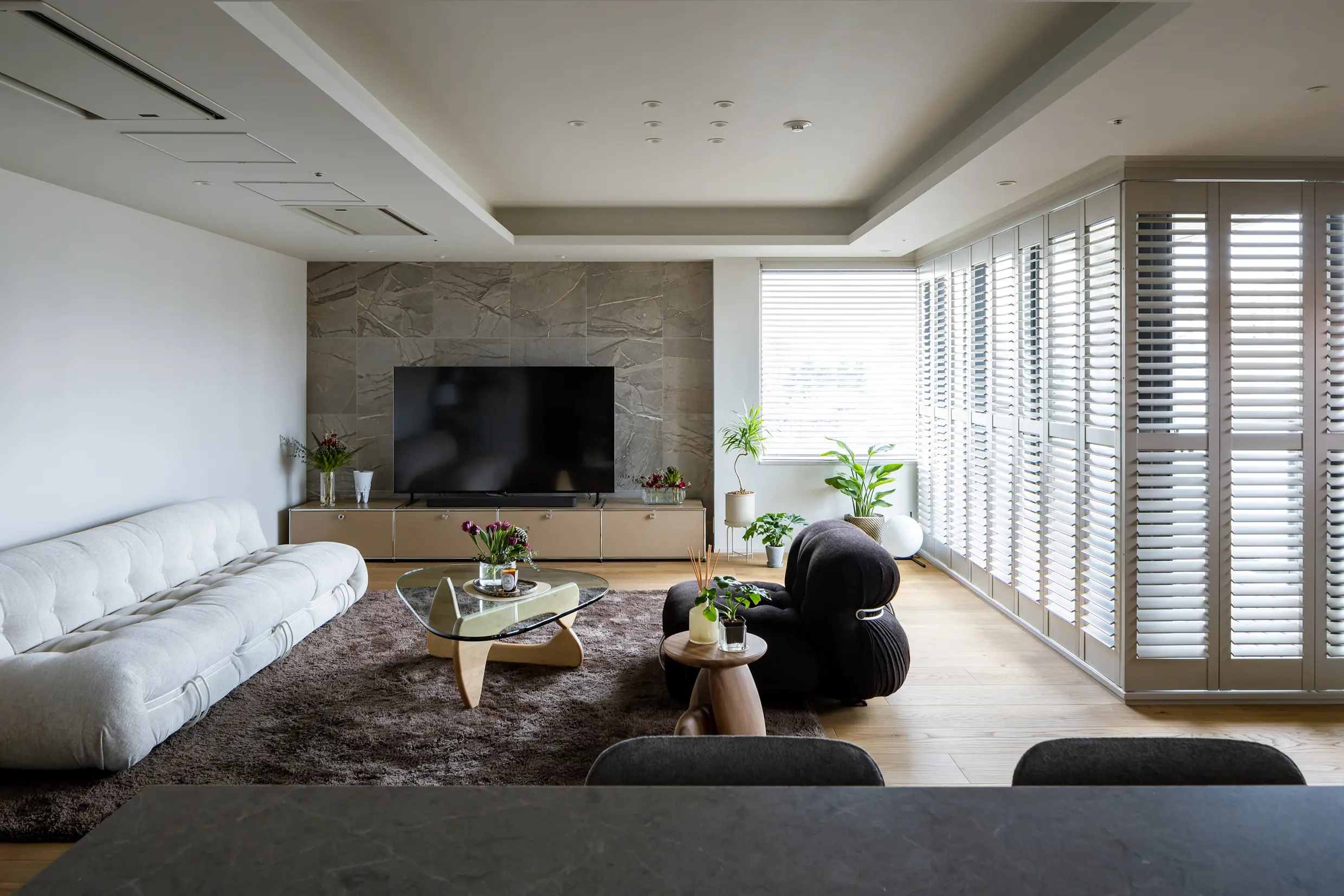 Open-plan living room with a wall-mounted TV on a marble accent wall, white sectional sofa, glass coffee table, and large windows with blinds.