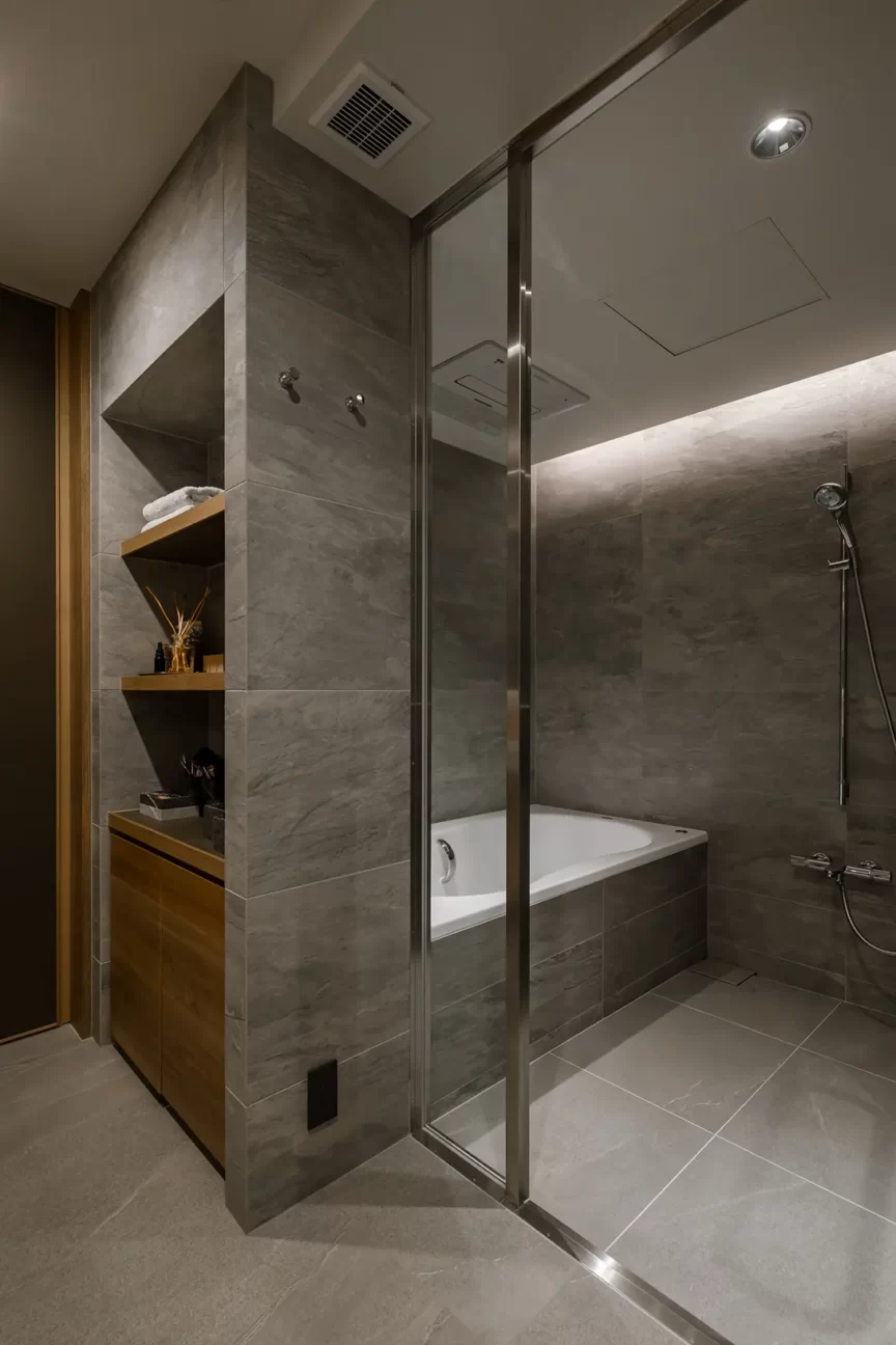 Luxurious gray-tiled bathroom with a glass shower enclosure and built-in wooden shelves beside a white bathtub.