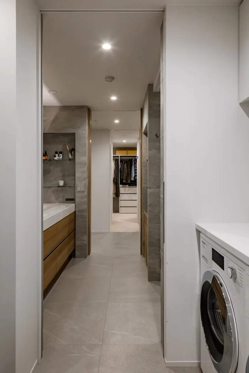 Narrow laundry hallway with a front-loading washing machine on the right and gray tiled floor, leading to a walk-in closet.