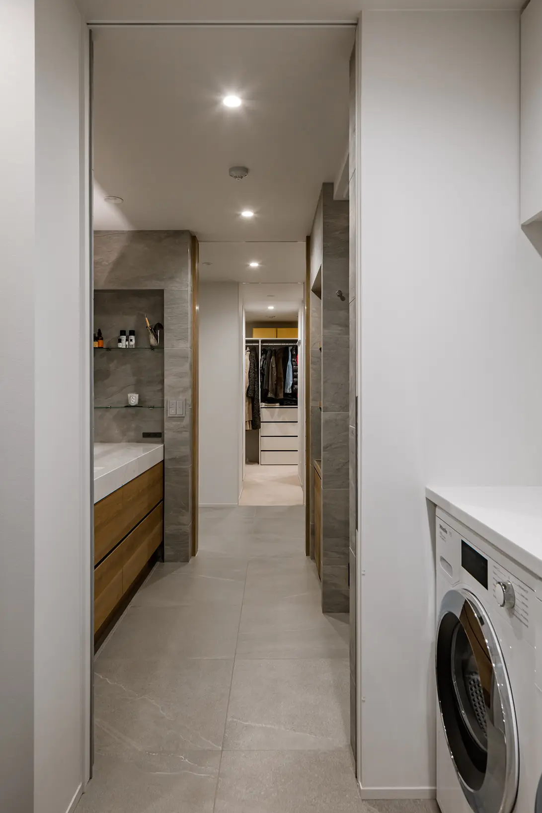 Narrow laundry hallway with a front-loading washing machine on the right and gray tiled floor, leading to a walk-in closet.