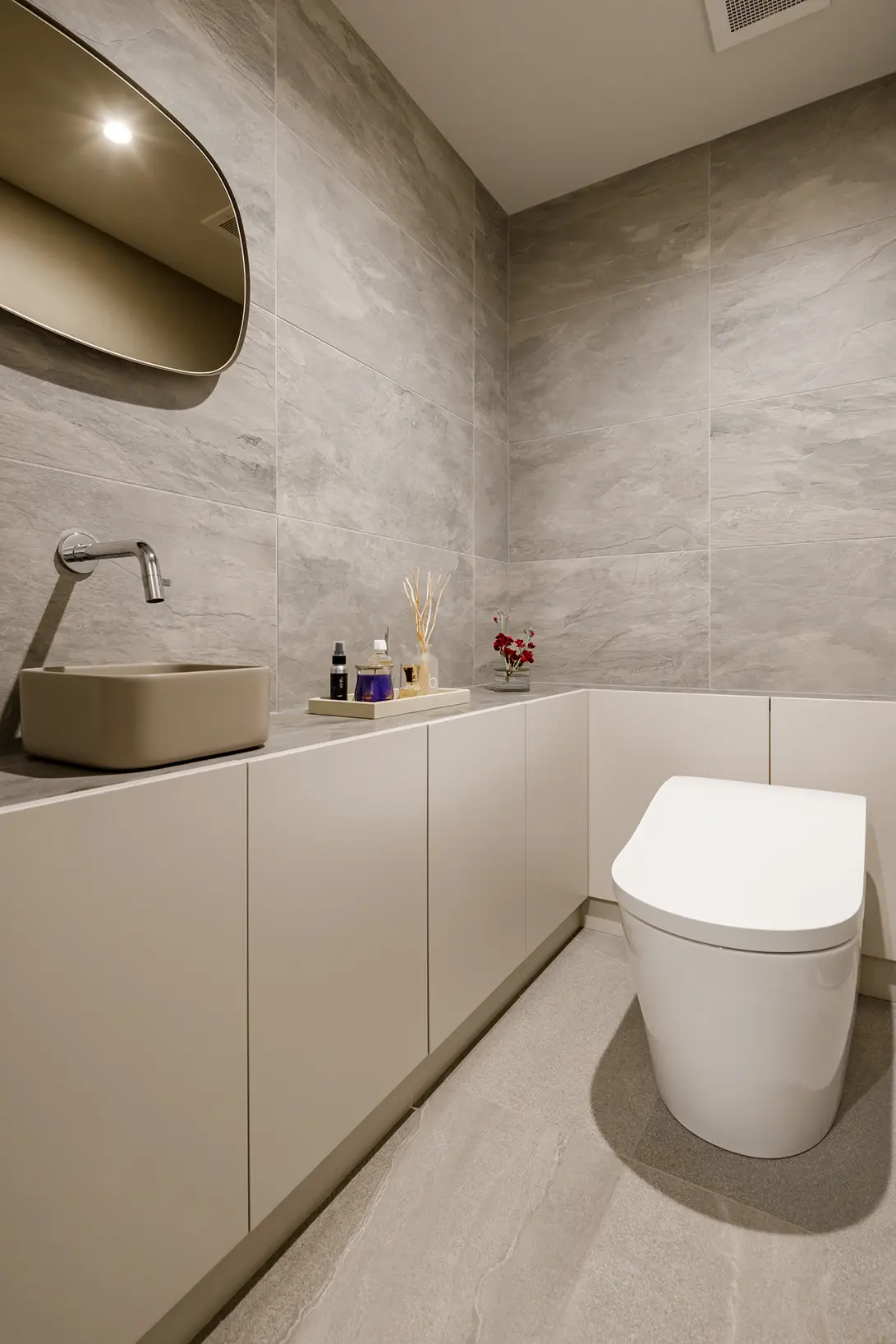 Minimalist bathroom with grey tiled walls, a beige rectangular sink, wall-mounted faucet, white cabinets, and a modern toilet.