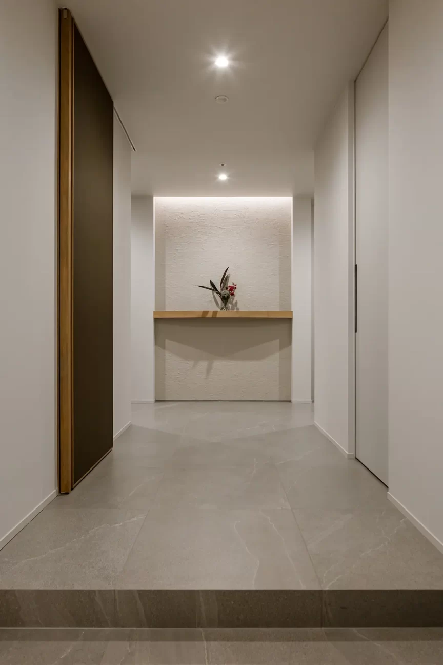 Narrow hallway leading to a minimalist reception alcove with a small vase of flowers on a light wood counter, soft recessed lighting