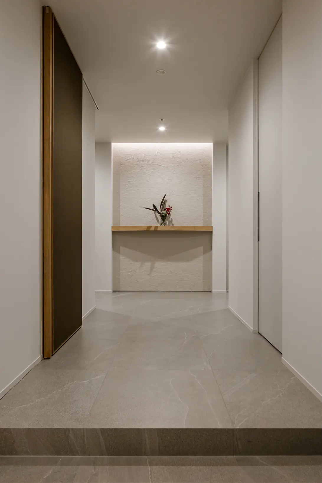 Narrow hallway leading to a minimalist reception alcove with a small vase of flowers on a light wood counter, soft recessed lighting