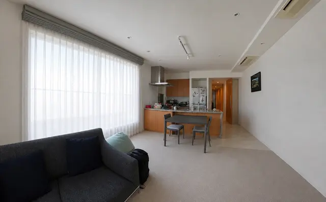 Bright open-plan living area with a gray sofa, large window with vertical blinds, and a kitchen island with two stools.