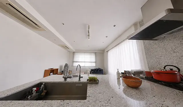 Open-plan kitchen with a granite island, double sink, and a red pot on the stove; living room visible in the background under bright light.
