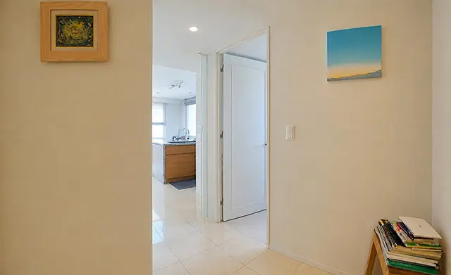 Bright hallway opening to a kitchen, with a small stack of books on a wooden stool and two framed paintings on the walls.