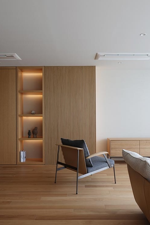 Minimalist living room featuring a vertical wood slat wall, built-in illuminated shelves, and a modern gray armchair on a light hardwood floor. The scene includes a low wooden cabinet along the right wall.