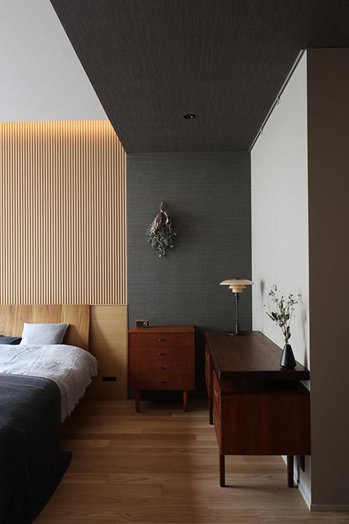 Modern bedroom with a slatted wooden wall behind the bed, a bedside dresser, and a desk with a lamp and vase; a dried bouquet hangs on a dark accent wall.