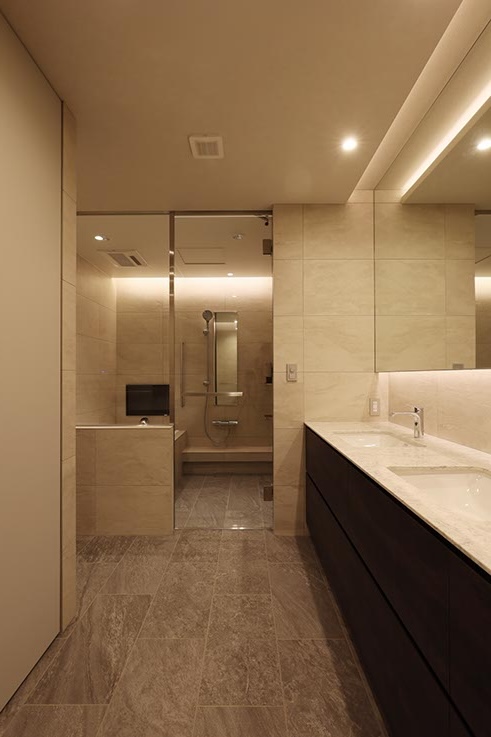 Modern beige-tiled bathroom with a long dark wood vanity and marble countertop on the right, and a walk-in shower at the far end