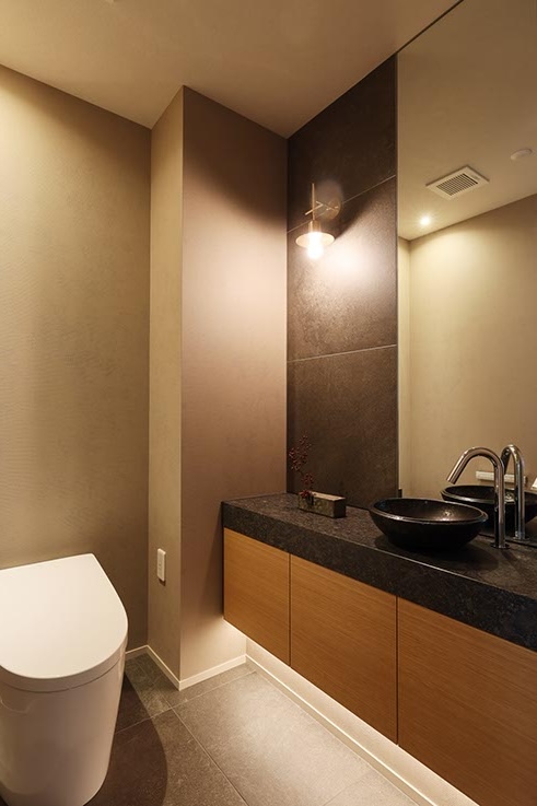 Contemporary bathroom with a white toilet on the left, dark stone countertop, and a black vessel sink atop wooden cabinets under warm lighting. a tall mirror reflects the space.