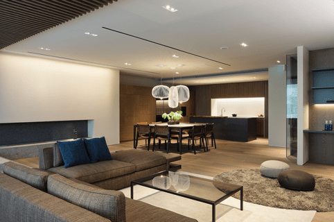 Open-concept living and dining area with a gray sectional sofa, glass coffee table, dining table, and a modern kitchen in the background.
