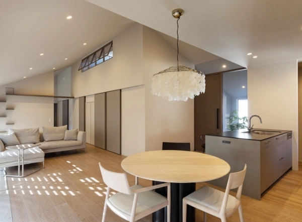 Open-plan living space with a round wooden dining table and white chairs, a sculpted chandelier, a modern kitchen island, and a gray sofa in the background.