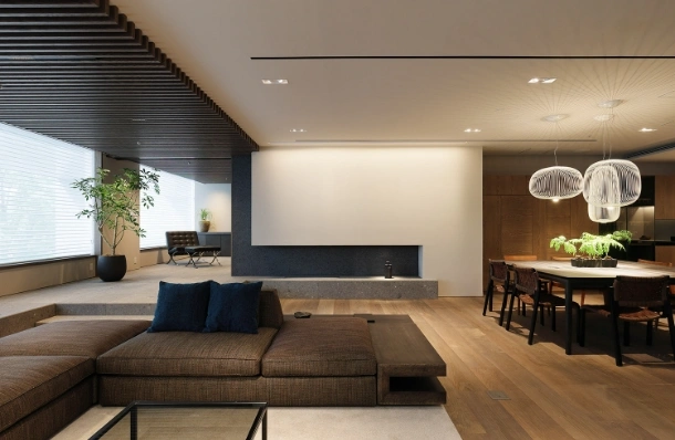 Open-plan living and dining area with a brown sectional sofa, blue cushions, a built-in fireplace wall, and a wooden floor beside a dining table with pendant lights.