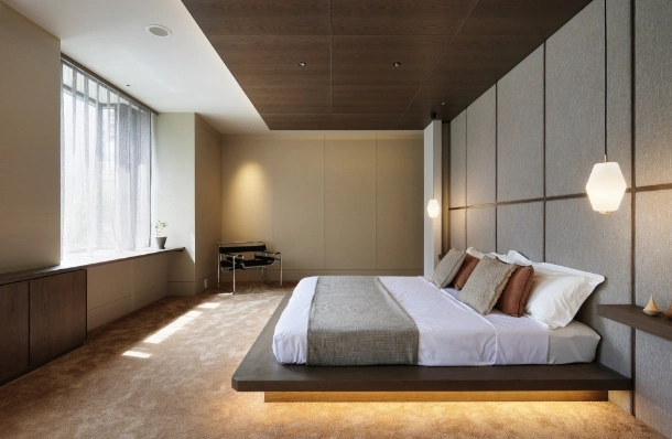Modern hotel bedroom with a low platform bed, padded grey headboard wall, and warm pendant lights; window seat and desk at the far end.