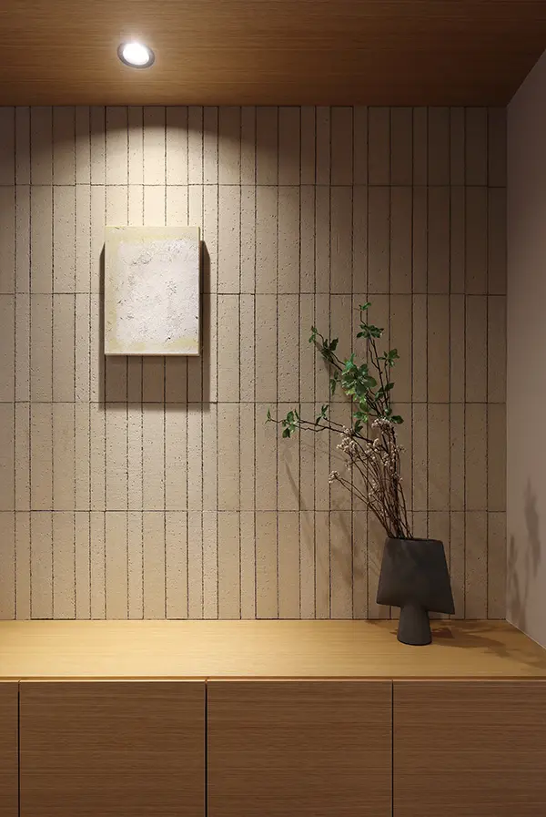 Minimalist interior corner: tiled wall, spotlight, small abstract painting, wooden countertop, and a dark vase with branches.