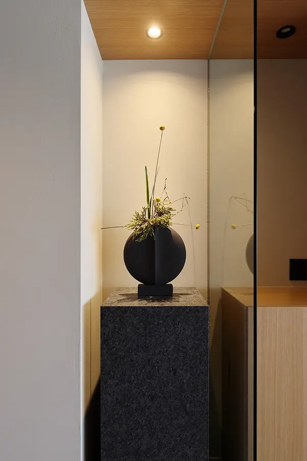 Contemporary interior display: a round black vase with dried stems on a dark granite pedestal in a narrow alcove under warm ceiling light.