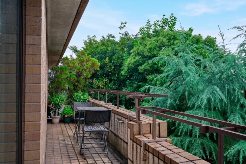 Balcony seating area with wicker chairs and a table, potted plants, and a dense green garden beyond the brick railing.