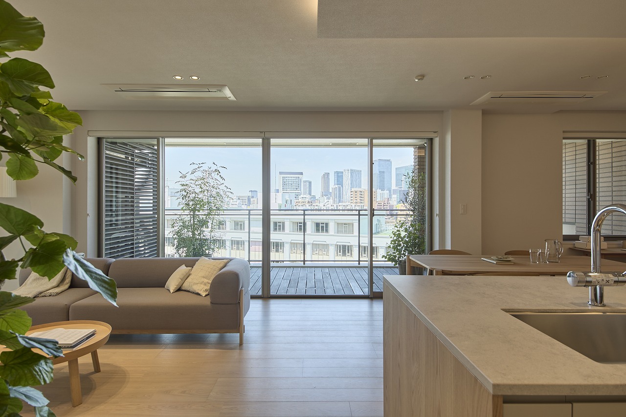 Bright living room with a gray sofa facing a glass sliding door to a balcony, overlooking a city skyline.