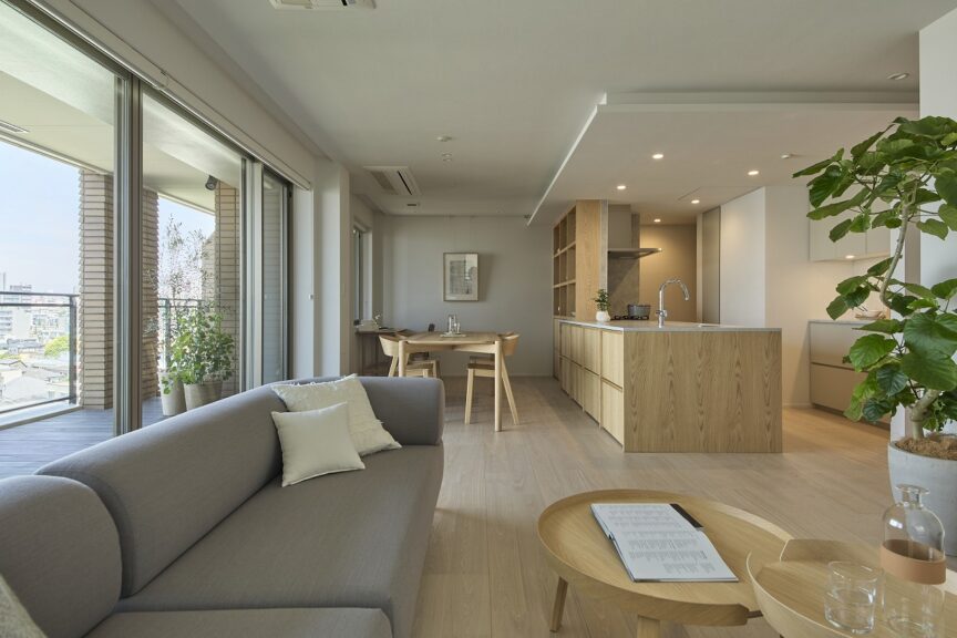 Bright, modern open-plan living area with a wooden kitchen island, gray sofa, and round coffee table near large windows and a balcony plant.