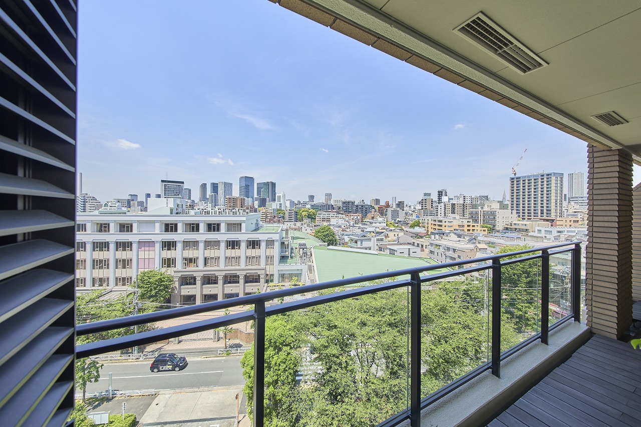 City skyline seen from a balcony with a glass railing, blue sky above, and a street with trees below.