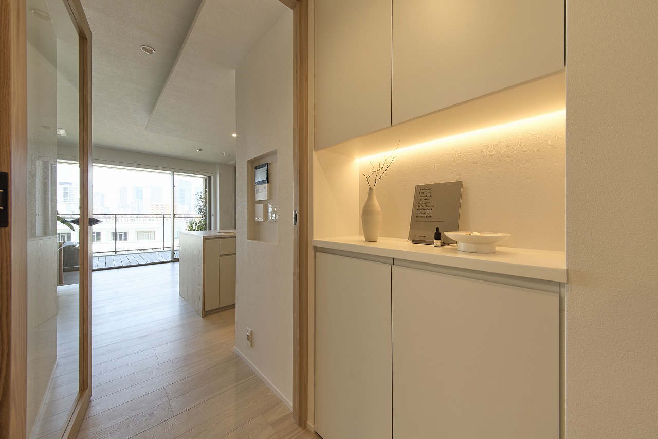 Bright, modern hallway leading to a balcony with city view; cream cabinets with warm under-cabinet lighting and a vase on the shelf.