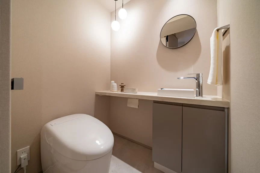 Modern beige bathroom with a rounded white toilet, floating vanity, and circular wall mirror with pendant lights above.