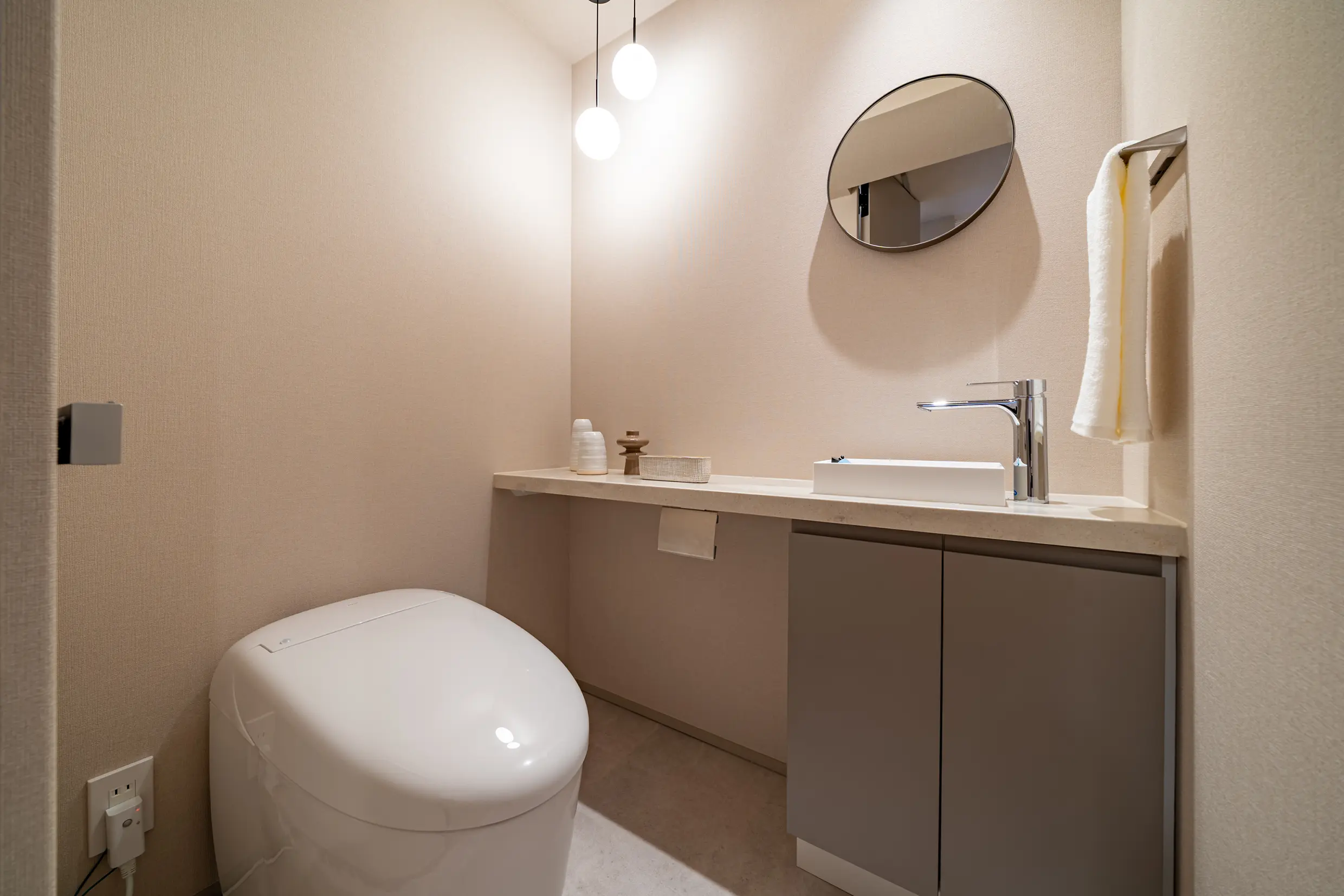 Modern beige bathroom with a rounded white toilet, floating vanity, and circular wall mirror with pendant lights above.