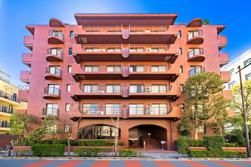 Six-story red brick apartment building with rounded balconies on both sides and a central ground-floor archway, set against a bright blue sky with trees in front.
