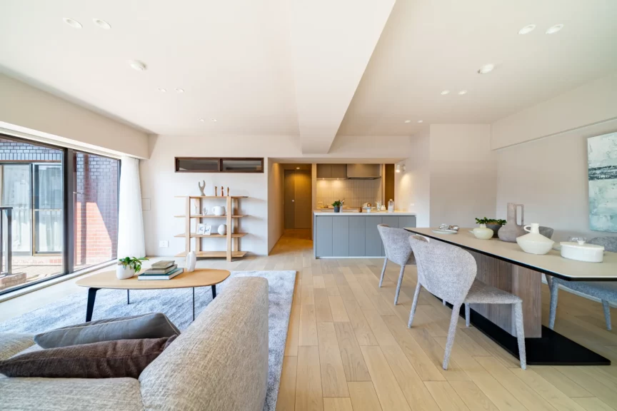 Open-plan living and dining space with light wood floors, grey sofas, a wooden bookshelf, and a modern kitchen island in the background.