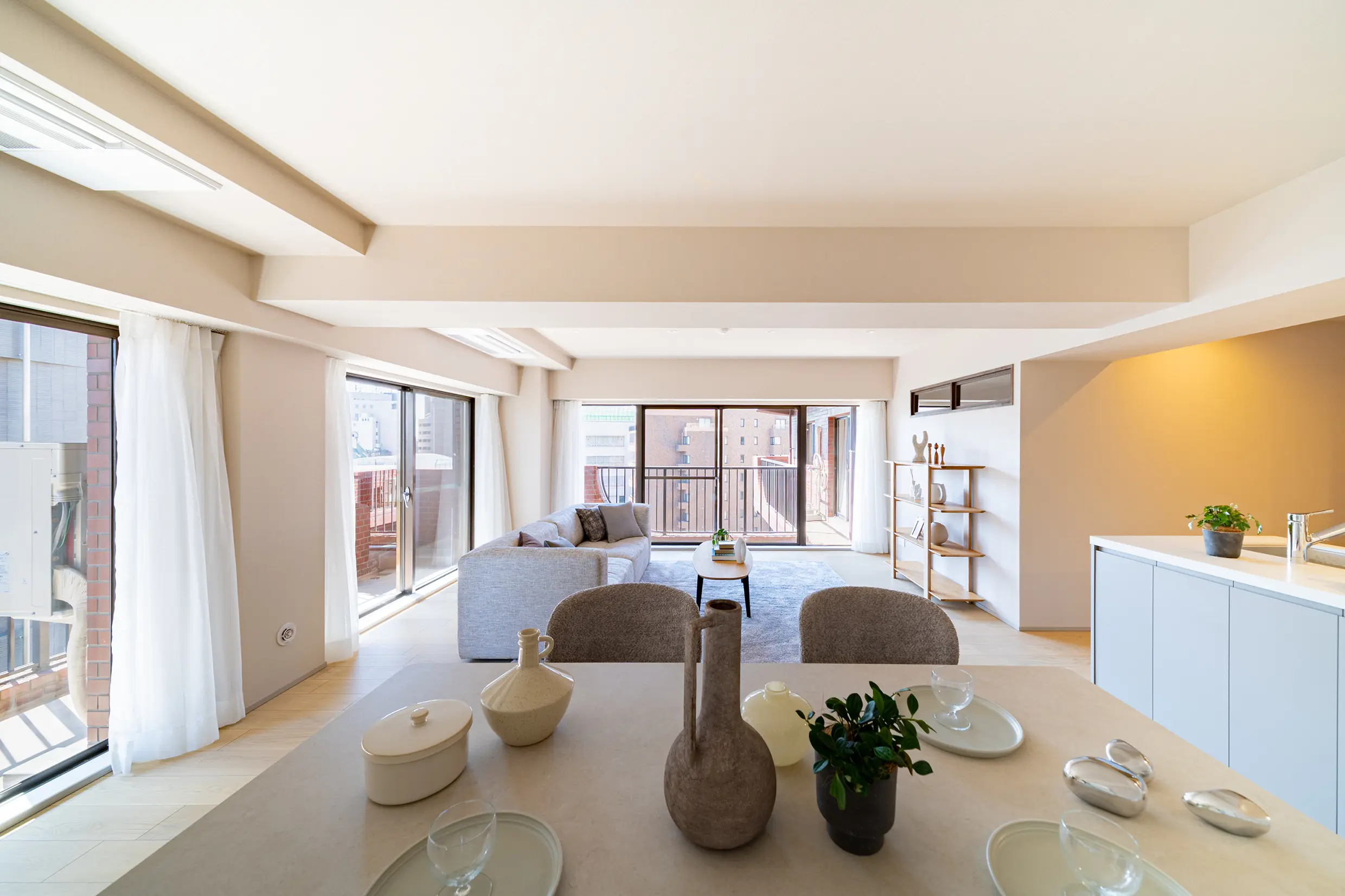 Bright open-plan living and dining area with large windows and white curtains, beige walls, and modern furniture.