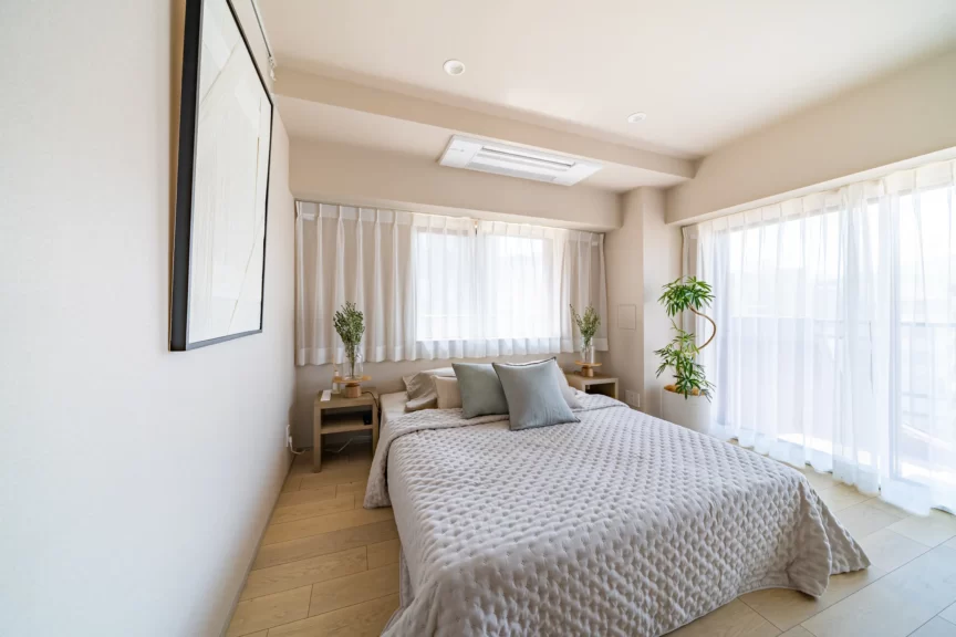 Bright neutral-toned bedroom with a large quilted gray bed, two small wood nightstands with plants, and sheer white curtains along tall windows.