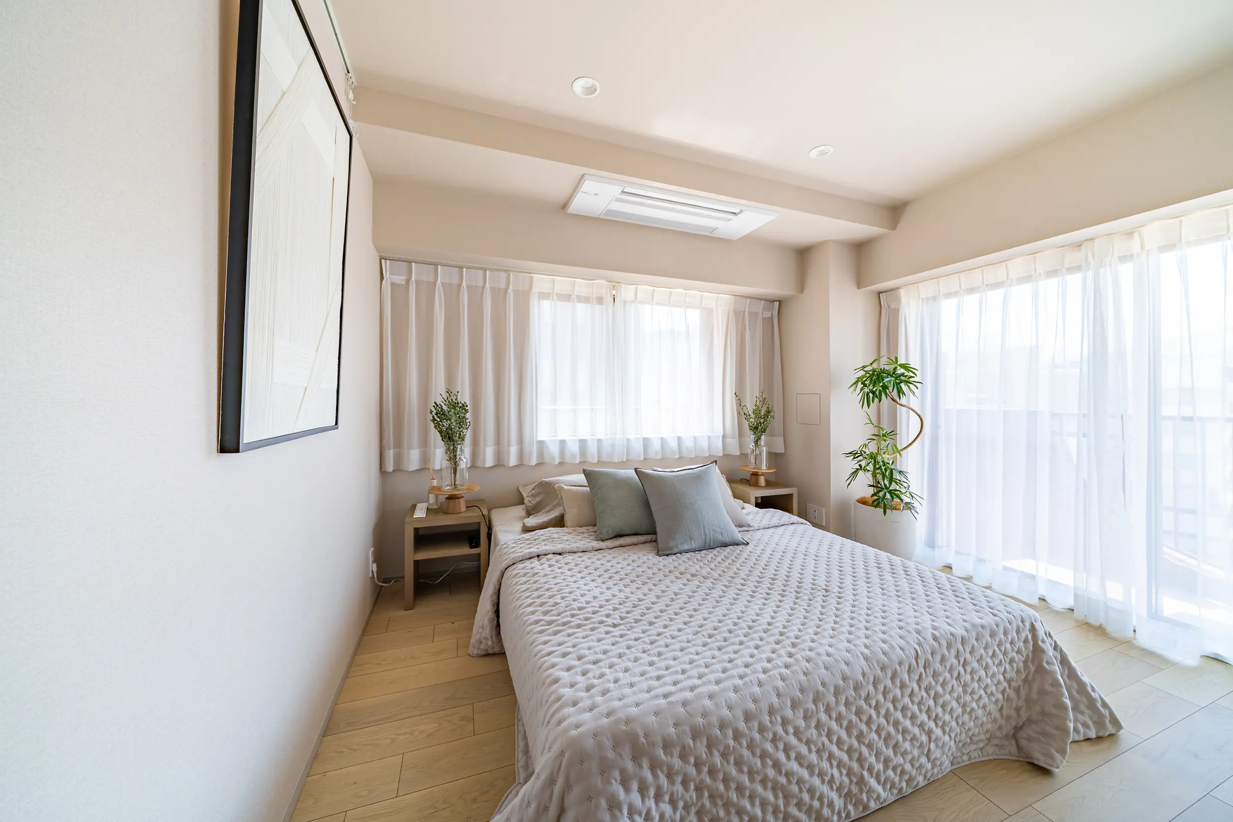 Bright neutral-toned bedroom with a large quilted gray bed, two small wood nightstands with plants, and sheer white curtains along tall windows.