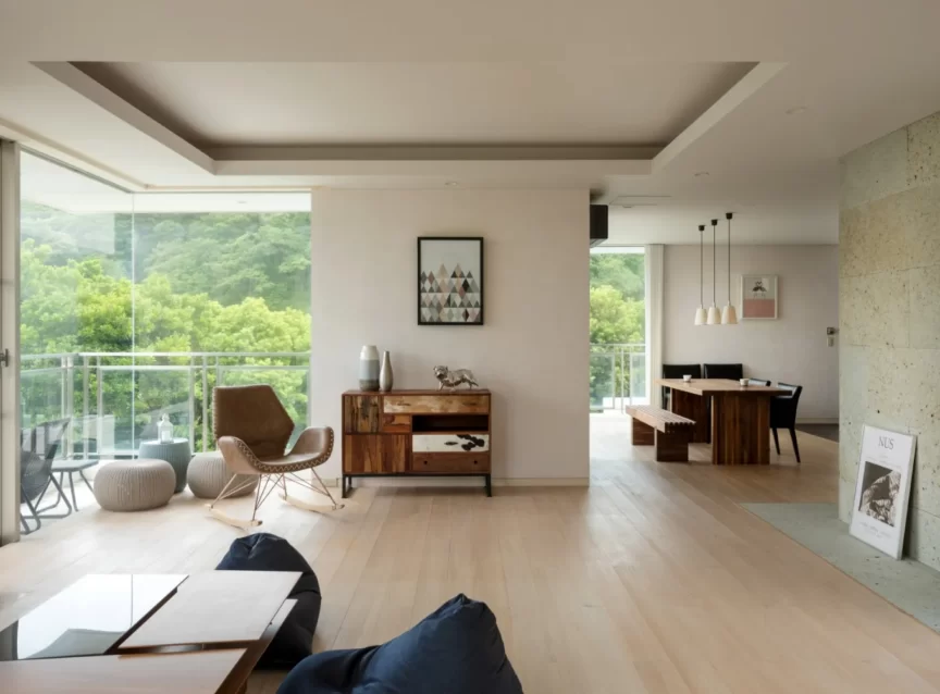 Bright open-plan living area with floor-to-ceiling windows showing green trees, light wood floors, and contemporary furnishings including a brown leather chair and wooden sideboard on the left. A wooden dining table with benches sits in the background near pendant lights.