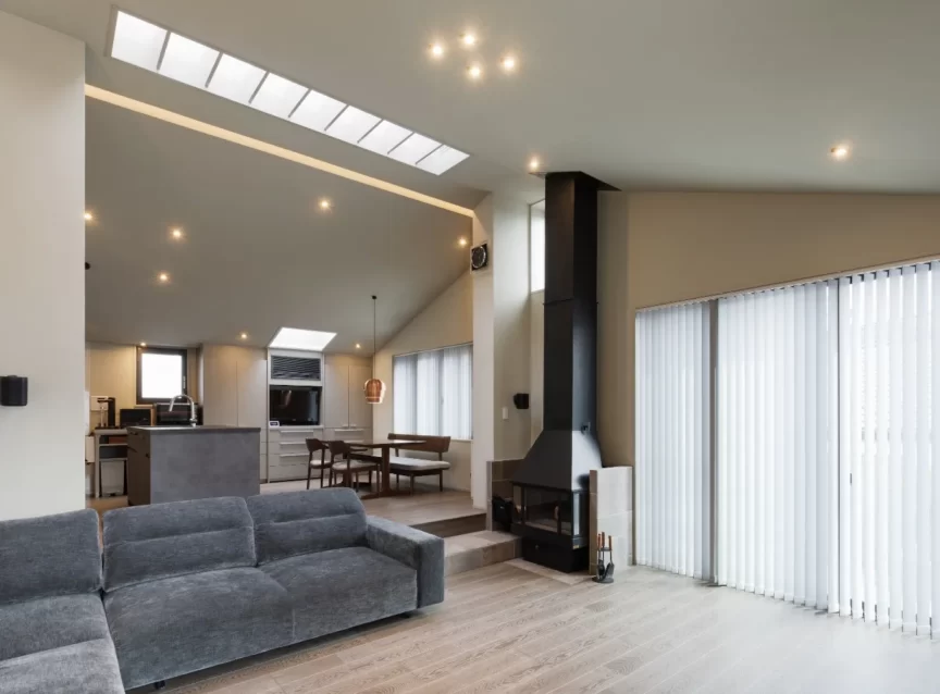 Open-concept living space with a gray sectional, kitchen island, dining table, and a tall black fireplace near large windows with vertical blinds.