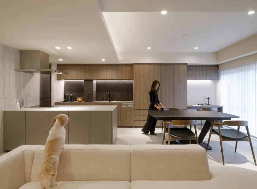 Open-plan kitchen with a beige island and wooden cabinets; a woman stands at a dark dining table while a small dog sits on a light sofa in the foreground.