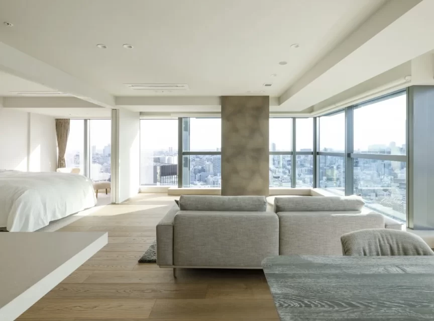 Spacious, modern living area in a high-rise apartment with large windows and a city view; gray sofa and dining table visible, bed area to the left.